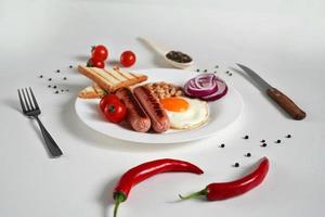 Traditional English Breakfast. Plate with Fried egg, two fried sausages, grilled bread toast, canned beans, blue onions and cherry tomatoes on a White Background. Copy space photo