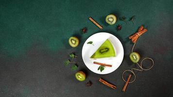 Homemade Cheesecake with Kiwi in a white plate on a Green background. Decoreted with slice of kiwi, cinnamon stick and leaves mint. Top view photo