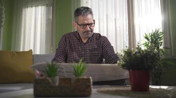 Serious and focused mature man with stern face reading newspaper at home. video