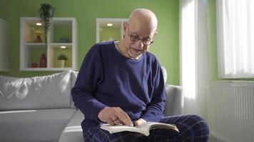 velho homem sentado em a sofá às casa de a janela, lendo uma livro. a velho homem é lendo dele livro às casa sozinho dentro uma ensolarado dia e é feliz. video