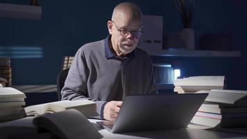 pensativo antiguo investigador hombre utilizando ordenador portátil en su oficina, transferir importante información él leer en libro a ordenador portátil.