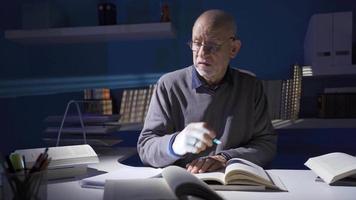 el antiguo hombre leyendo un libro, aprendizaje conocimiento, haciendo investigación es considerado. antiguo hombre hombre con lentes es sentado en el brillante estudiar habitación, leyendo un libro, preparando un tesis, haciendo investigación. video