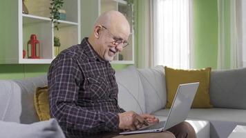 antiguo hombre sentado en sofá a hogar utilizando ordenador portátil divertido y contento. antiguo hombre a hogar utilizando ordenador portátil y sonriente, teniendo divertido y alegre. video
