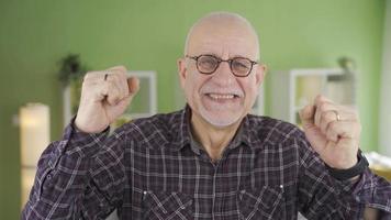 Cheerful old man rejoices at home looking at camera. Cute and happy old man looking at camera at home rejoicing at the good news he has received. video