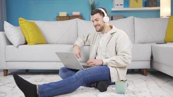 contento joven hombre buscando en línea desde computadora solo a hogar. joven hombre utilizando su ordenador portátil búsquedas en línea con auriculares. video