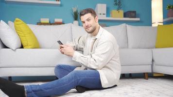 giovane uomo seduta solo a casa guardare a Telefono e sorridente a telecamera. giovane uomo sorridente a telecamera mentre giocherellare con Telefono e dando un' positivo gesto con il suo mano. video