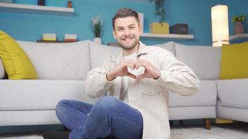 Man sitting on floor and making heart sign towards camera. Young man smiling and looking at camera with hands up. video