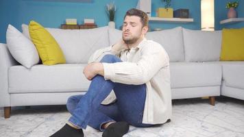 The sad young man sits alone in the living room and thinks about his experiences. Young depressed man sitting alone on the floor thinks about what has happened to him and sighs. video