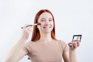 alegre pelirrojo joven mujer utilizando maquillaje cepillo y colorete aislado en blanco antecedentes foto