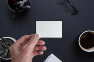 Blank business card in hand mockup. Office desk in background with plant, coffee, glasses, pens, pad photo