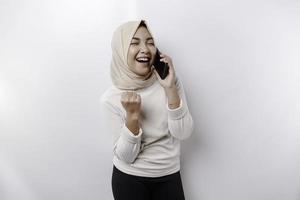 un contento asiático musulmán mujer vistiendo un Pañuelo, participación su teléfono, aislado por blanco antecedentes foto