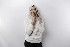 un pensativo joven asiático musulmán mujer vistiendo un hijab y participación su barbilla mientras hablando en el teléfono aislado por un blanco antecedentes foto