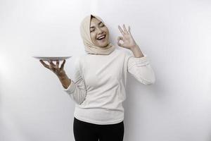 A smiling Asian Muslim woman is fasting and hungry and holding and pointing to a plate photo