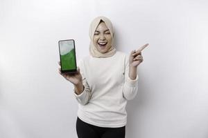 Excited Asian Muslim woman wearing headscarf pointing at the copy space beside her while showing her phone, isolated by white background photo