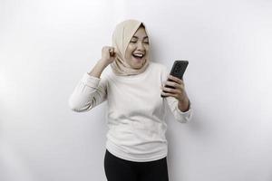 A young Asian Muslim woman with a happy successful expression wearing a hijab and holding smartphone isolated by white background photo