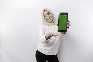 A happy Asian Muslim woman wearing a headscarf, showing her phone screen, isolated by white background photo