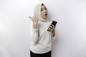 Shocked Asian Muslim woman wearing hijab pointing at the copy space beside her while holding her phone, isolated by white background photo