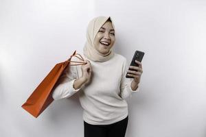 Portrait Asian Muslim woman standing excited holding an online shopping bag and her smartphone, studio shot isolated on white background photo