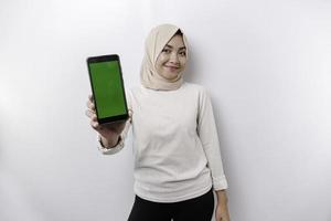 A happy Asian Muslim woman wearing a headscarf, showing her phone screen, isolated by white background photo