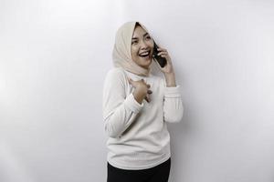 A happy Asian Muslim woman wearing a headscarf, holding her phone, isolated by white background photo