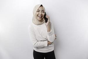 A happy Asian Muslim woman wearing a headscarf, holding her phone, isolated by white background photo