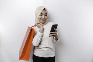 Portrait Asian Muslim woman standing excited holding an online shopping bag and her smartphone, studio shot isolated on white background photo