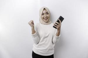 A young Asian Muslim woman with a happy successful expression wearing a hijab and holding smartphone isolated by white background photo