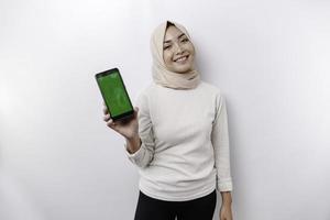 A happy Asian Muslim woman wearing a headscarf, showing her phone screen, isolated by white background photo