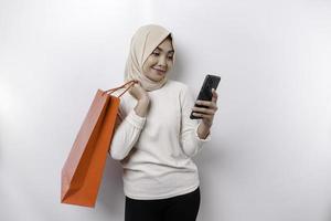 Portrait Asian Muslim woman standing excited holding an online shopping bag and her smartphone, studio shot isolated on white background photo