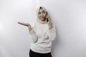 un pensativo joven asiático musulmán mujer vistiendo un hijab y señalando Copiar espacio junto a su mientras hablando en el teléfono aislado por un blanco antecedentes foto