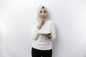 An Asian Muslim woman is fasting and hungry and holding a plate while looking aside thinking about what to eat photo