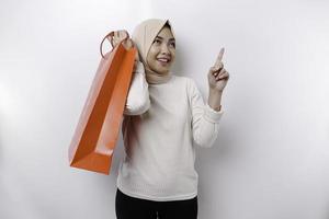 A happy Asian Muslim woman standing excited holding a shopping bag and pointing to copy space above her isolated on white background photo