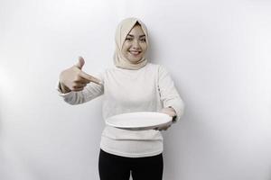 A smiling Asian Muslim woman is fasting and hungry and holding and pointing to a plate photo