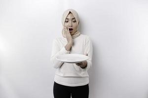 A shocked Asian Muslim woman is fasting and hungry and holding a plate with her mouth wide open photo