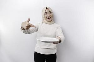 A smiling Asian Muslim woman is fasting and hungry and holding and pointing to a plate photo