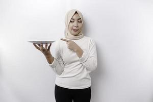 A smiling Asian Muslim woman is fasting and hungry and holding and pointing to a plate photo