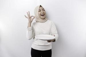 A smiling Asian Muslim woman is fasting and hungry and holding and pointing to a plate photo