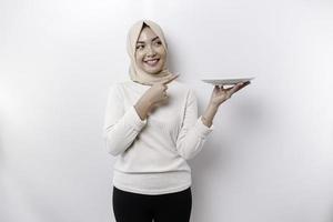 A smiling Asian Muslim woman is fasting and hungry and holding and pointing to a plate photo