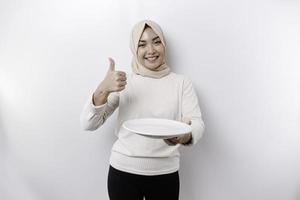 A smiling Asian Muslim woman is fasting and hungry and holding and pointing to a plate photo