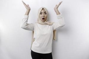 Shocked Asian Muslim woman wearing a headscarf pointing at the copy space on top of her, isolated by a white background photo