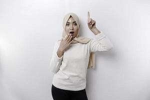 Shocked Asian Muslim woman wearing a headscarf pointing at the copy space on top of her, isolated by a white background photo