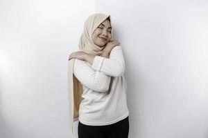 Young beautiful Asian Muslim woman wearing a headscarf over white background hugging herself happy and positive, smiling confident. Self-love and self-care photo