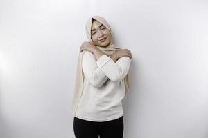 joven hermosa asiático musulmán mujer vistiendo un Pañuelo terminado blanco antecedentes abrazando sí misma contento y positivo, sonriente seguro. amor propio y cuidados personales foto