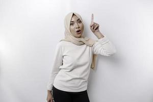 Shocked Asian Muslim woman wearing a headscarf pointing at the copy space on top of her, isolated by a white background photo