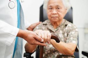 Doctor help Asian elderly disability woman patient holding walking stick in wrinkled hand at hospital. photo