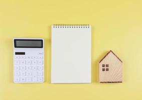 plano diseño de de madera casa modelo, blanco calculadora, blanco página abrió cuaderno en amarillo antecedentes con Copiar espacio, hogar compra concepto. foto