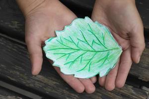 un verde hoja dibujado con de colores lapices es pulcramente retenida en el manos de un pequeño niño. foto