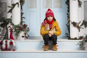 Handsome little boy in christmas. Funny child holds old skates and sits on the porch of a house decorated with Christmas tree decorations photo