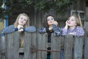 Three funny village girls are standing near the old village fence. Bored country girls. photo