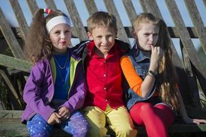 Funny little children girls with bright makeup dressed in the style of the nineties and a boy in a red shirt are sitting on a bench by a skewed fence. Russian village children. photo